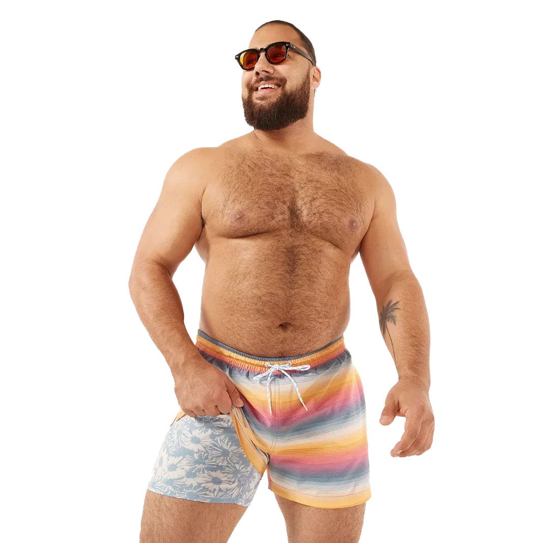 A man wearing multicolored striped swim shorts with blue floral design on one side, sunglasses, and displaying a tattoo.