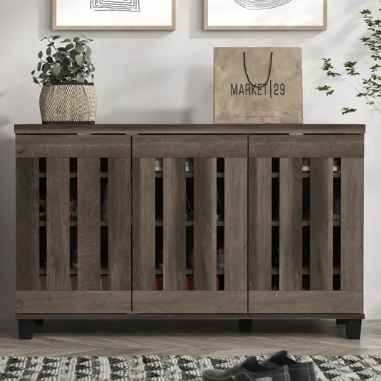 Brown wooden shoe cabinet with slatted doors, topped with a potted plant and a shopping bag labeled \“Market 29\“. Two abstract framed artworks hang above, and there's a patterned rug and shoes on the floor in front.