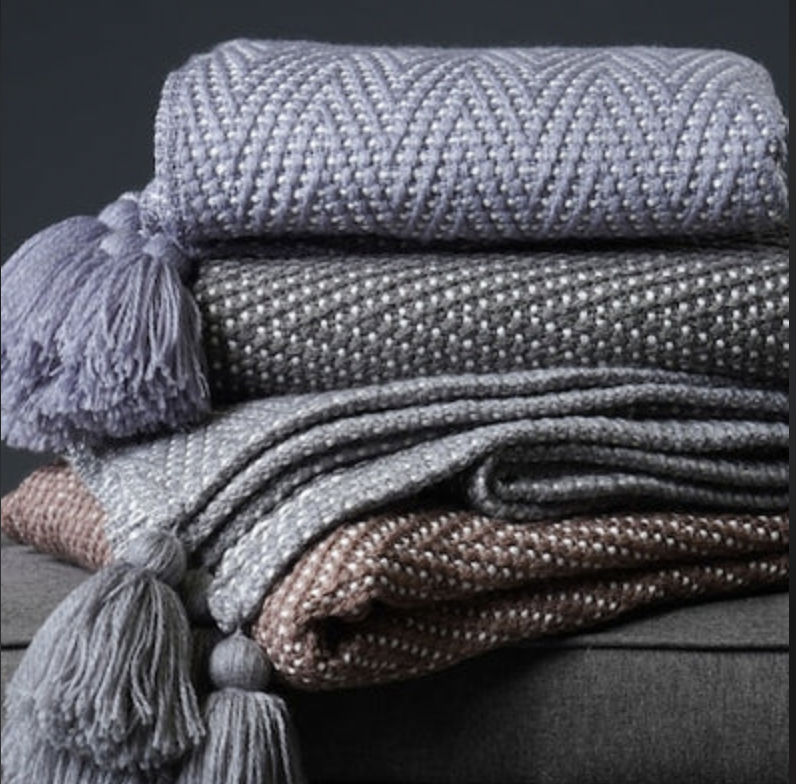Three knitted throw blankets in shades of purple and gray, stacked on top of each other with tassels on the corners.
