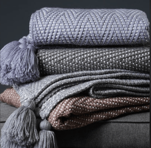 Three knitted throw blankets in shades of purple and gray, stacked on top of each other with tassels on the corners.