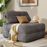 A gray corduroy armchair with a backrest is adorned with a brown cushion and a beige patterned throw. It is situated next to a small lamp and a plant, with abstract wall art in the background.