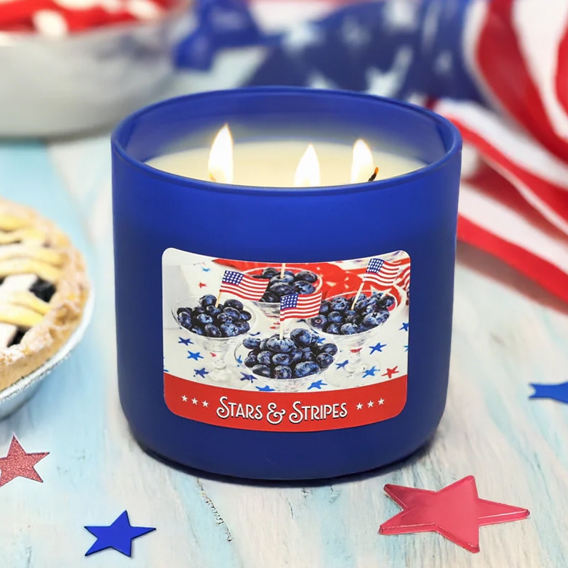 A blue candle jar labeled \“Stars & Stripes,\“ with three wicks, featuring blueberries and small American flags. The setting includes scattered star decorations and a pie, suggesting a festive, patriotic theme.