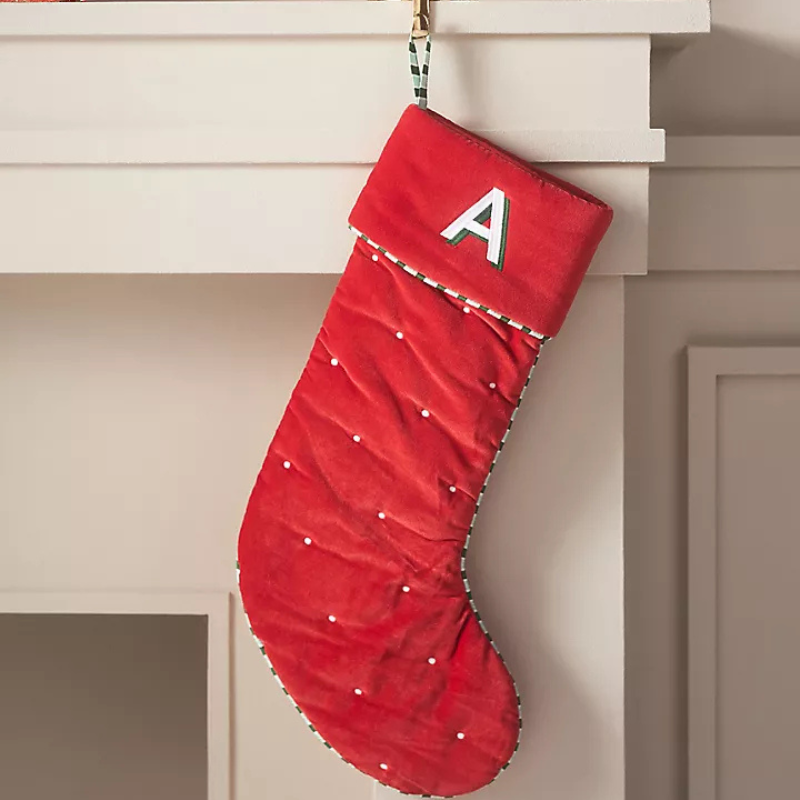 A red velvet stocking with white dots and a monogrammed \“A\“ on the cuff, trimmed with striped piping, hangs from a fireplace mantle.