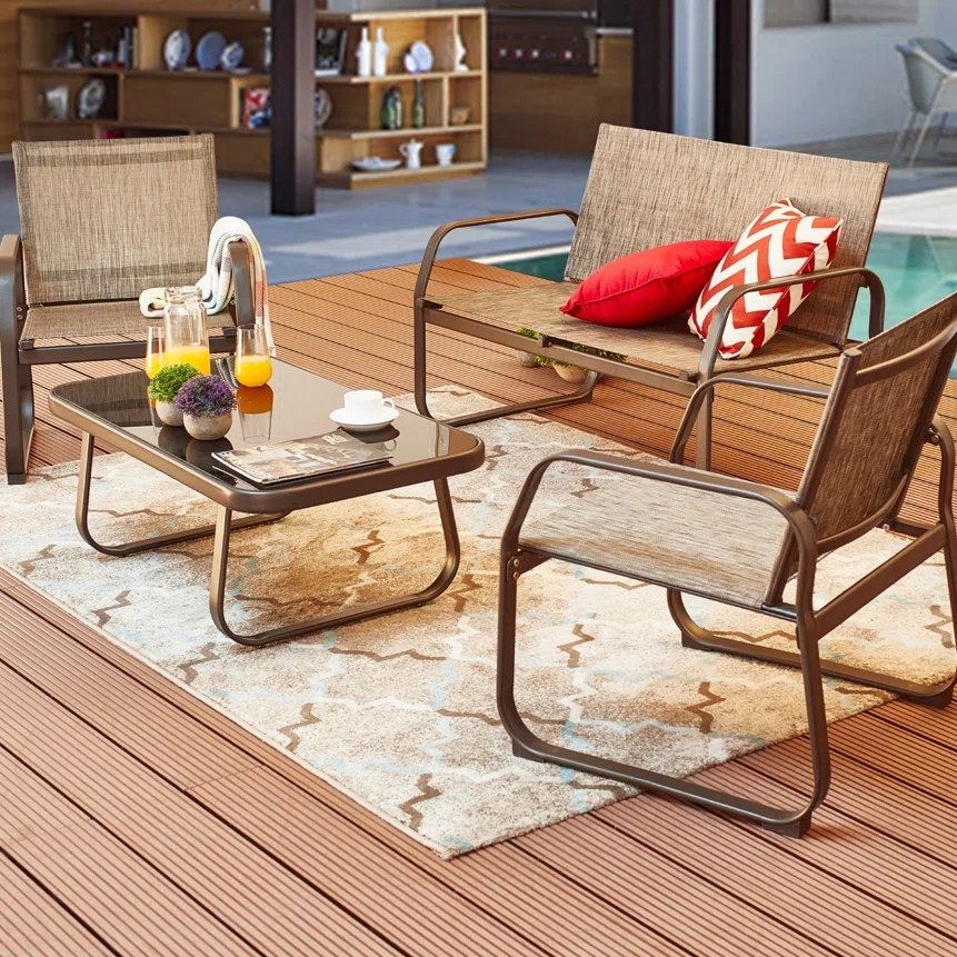 Outdoor patio set with two chairs, a loveseat, and a glass-top coffee table on a deck. The setup includes colorful cushions, a patterned rug, and a tray with drinks.