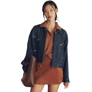 A woman is wearing a dark blue denim jacket over a brown collared sweater and a brown skirt. She is holding a large brown handbag.