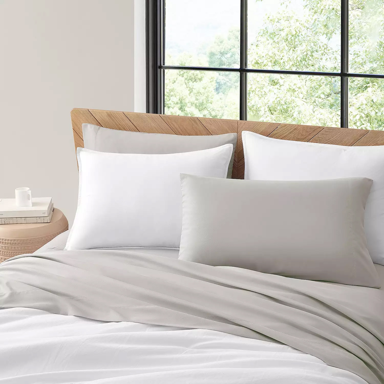 Koolaburra Temp-Control Sheets feature a soft gray color, offering a minimalist and modern look. The set includes two pillows and a cozy duvet, set against a light wood headboard and large window with green foliage outside.