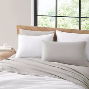 Koolaburra Temp-Control Sheets feature a soft gray color, offering a minimalist and modern look. The set includes two pillows and a cozy duvet, set against a light wood headboard and large window with green foliage outside.