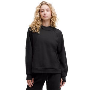 A woman is wearing a black, oversized lululemon hoodie with a relaxed fit, featuring a crew neckline and raglan sleeves. The hoodie includes a front pocket and is paired with matching black pants.