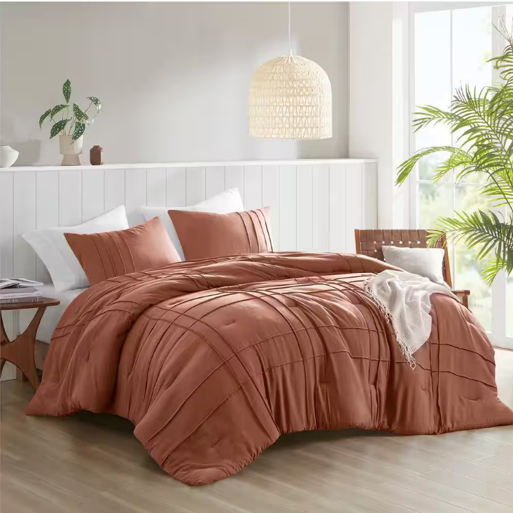 A rust-colored comforter set with matching pillow shams is on a bed, accompanied by white accent pillows. The room features a woven pendant light, potted plants, and a light wood nightstand.