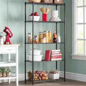 A black metal shelving unit holds kitchen items like bowls, jars filled with pasta, oils, and a basket of onions and potatoes. To the side, a countertop with a red mixer and nearby kitchen items is visible.