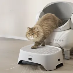 A fluffy cat steps off an automatic self-cleaning litter box onto a step platform. The platform has a brand name \“neakasa\“ and features control buttons on the front.