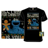 Black T-shirt featuring a festive design with a blue character holding a gingerbread cookie, surrounded by snowflakes, and the text \“All I Want for Christmas is You.\“