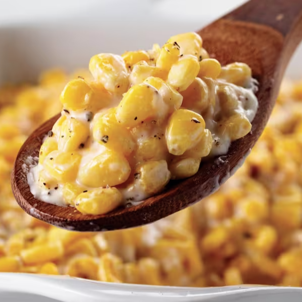 A wooden spoon holds creamy corn mixed with a sauce, garnished with black pepper. The dish is in a white serving pan.