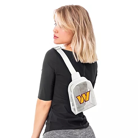 A person models a clear crossbody sling bag with a white strap and an orange and yellow logo, worn over a black shirt and gray leggings.