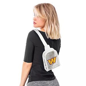 A person models a clear crossbody sling bag with a white strap and an orange and yellow logo, worn over a black shirt and gray leggings.
