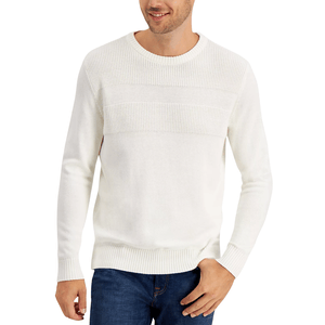 A man wearing a white knitted sweater with textured patterns across the chest and ribbed cuffs and hem, paired with dark blue jeans.