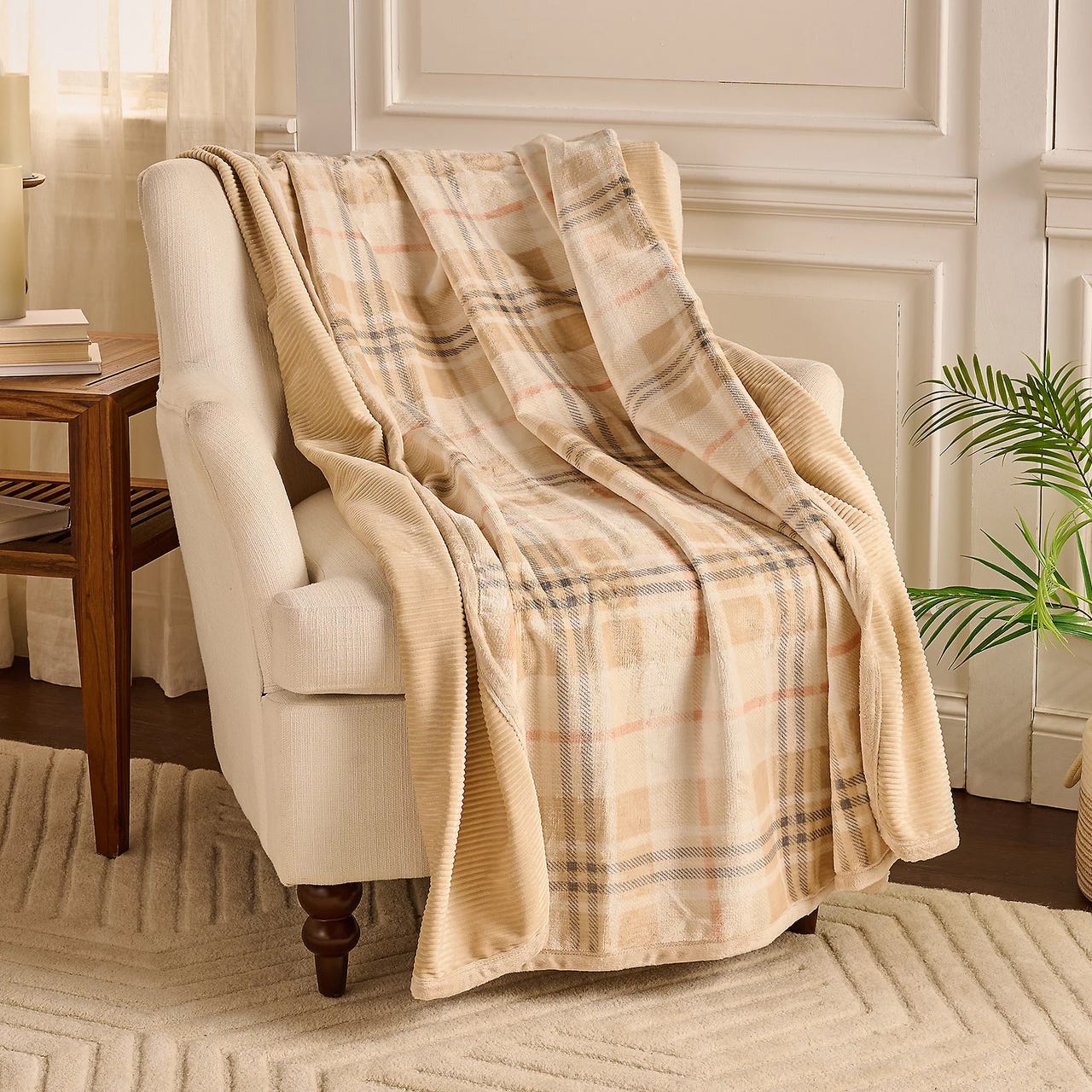 A plaid throw blanket with a beige, tan, and light pink checkered pattern is draped over a beige armchair, set on a textured light-colored rug, near a small wooden side table with books.