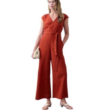 This is an image of a woman wearing an orange jumpsuit with a textured fabric and waist tie. She is holding a small woven handbag and wearing white sandals. The style is casual and chic.
