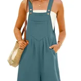 A woman is wearing teal short overalls with front buttons and pockets, paired with a white tube top. She carries a woven straw bag and wears a necklace.