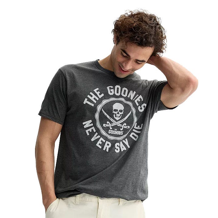 A person wearing a dark gray t-shirt featuring a skull and crossbones design with the text \“The Goonies Never Say Die.\“