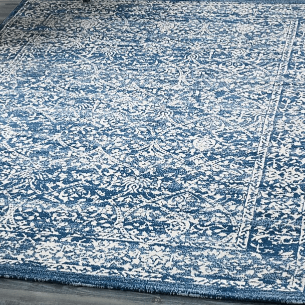 A blue and white area rug with an intricate pattern lies on a wooden floor. A chair with a cushion, a small table, and books on shelves are visible in the surrounding space.