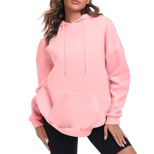 A woman is wearing a light pink oversized hoodie with a front pocket, drawstring hood, and long sleeves. The hoodie pairs with black shorts and features a relaxed, comfortable fit.