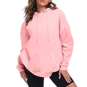 A woman is wearing a light pink oversized hoodie with a front pocket, drawstring hood, and long sleeves. The hoodie pairs with black shorts and features a relaxed, comfortable fit.