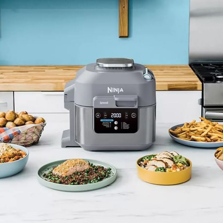 A Ninja brand kitchen appliance, possibly a multi-cooker, with cooked food displayed on plates nearby.