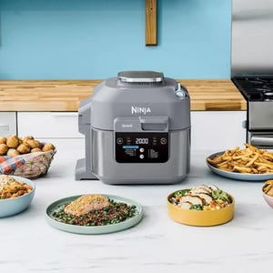 A Ninja brand kitchen appliance, possibly a multi-cooker, with cooked food displayed on plates nearby.