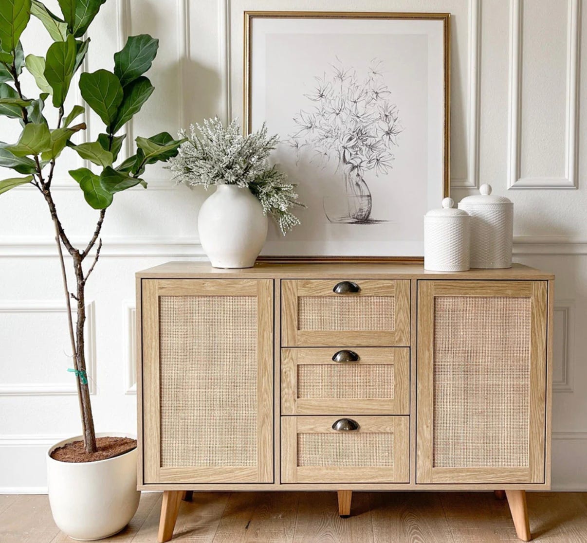 A wooden sideboard with rattan paneling accompanied by a potted plant, a white vase with artificial flowers, and framed sketch artwork.