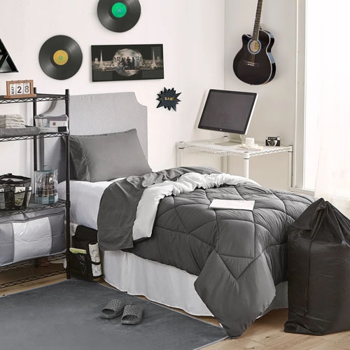 Bedroom setup with a twin bed featuring a gray comforter, a side shelving unit, wall-mounted records, a guitar, and a computer desk with a monitor. Slippers and a laundry bag are on the floor, with a gray rug.