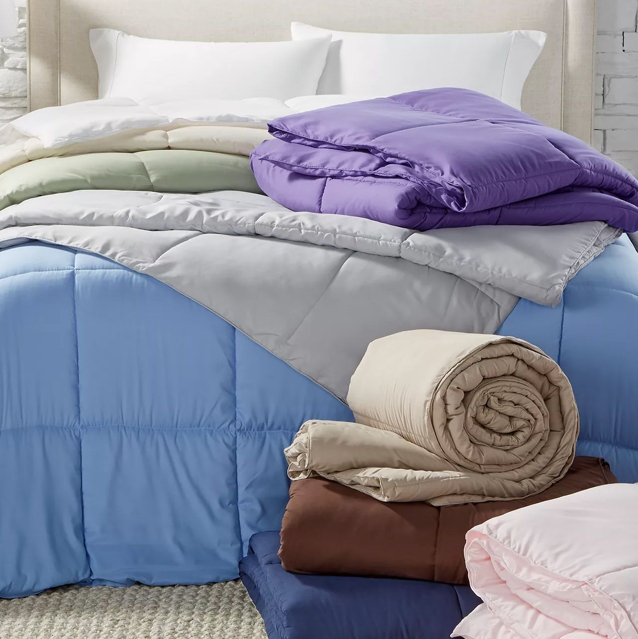 Multiple folded and rolled comforters in various colors, including blue, purple, beige, brown, and pink, are stacked on a bed with white pillows, creating a cozy and colorful display.