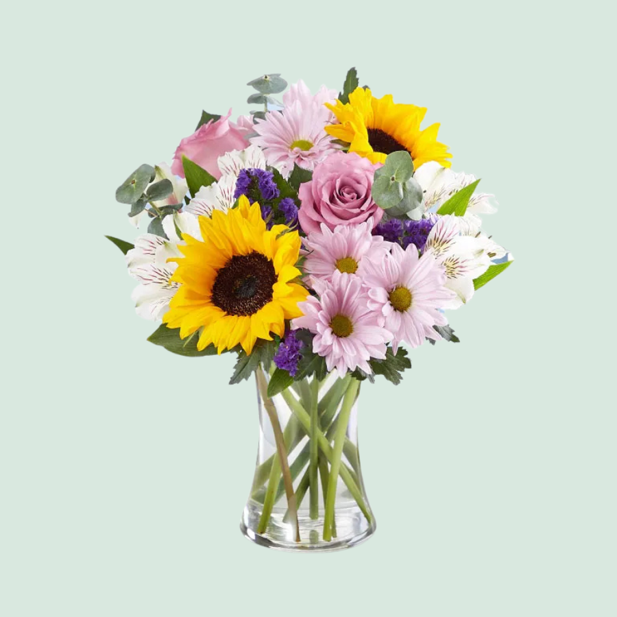 A bouquet featuring sunflowers, pink roses, alstroemeria, and pale purple chrysanthemums in a clear glass vase, accented with purple and green foliage.