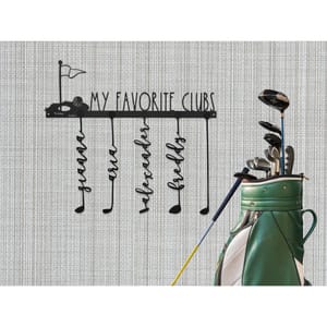 A metal wall sign features the text \“My Favorite Clubs\“ with a golf cart and flag design. Below are hooks shaped like golf clubs with names: guama, rose, alexander, and freddy. A green golf bag with various clubs stands beside the sign.