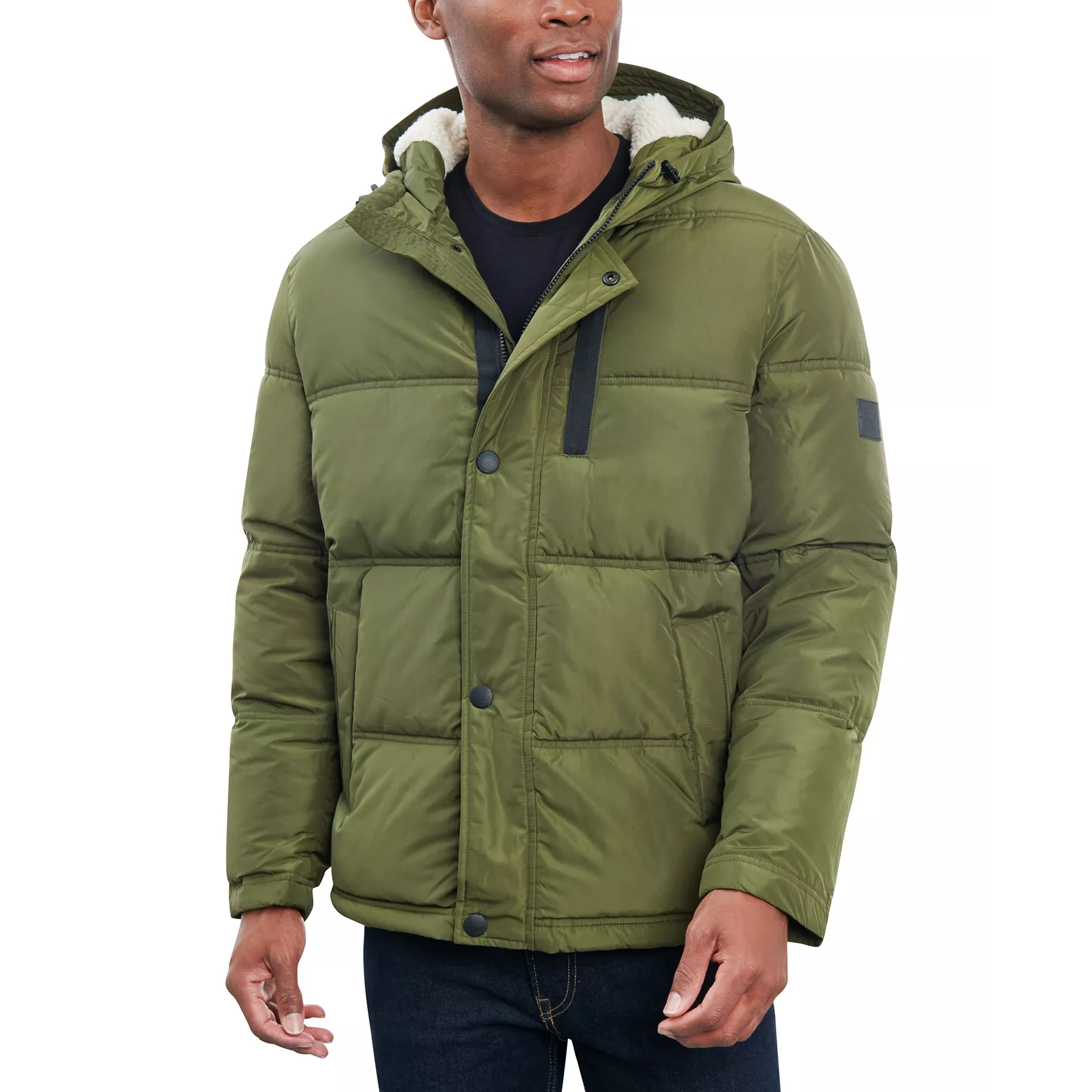 A man wearing an olive green puffer jacket with a hood, black buttons, and a sherpa lining, paired with dark jeans.
