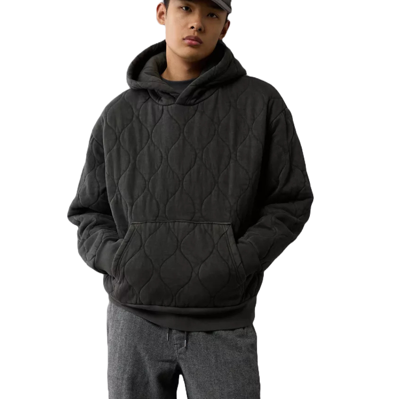 A dark, heavyweight quilted hoodie featuring a textured pattern, kangaroo pocket, and drawstring hood, paired with gray trousers and a cap.