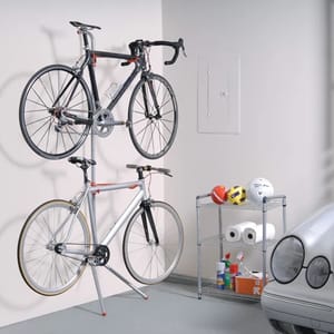 Two bicycles are mounted on a vertical bike rack against a wall, with additional storage space on a small metal shelf unit containing helmets and cleaning supplies.