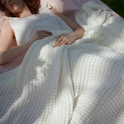 A woman is lying on her back outdoors, covered to her chest with a textured queen-sized blanket in a soft, light color.