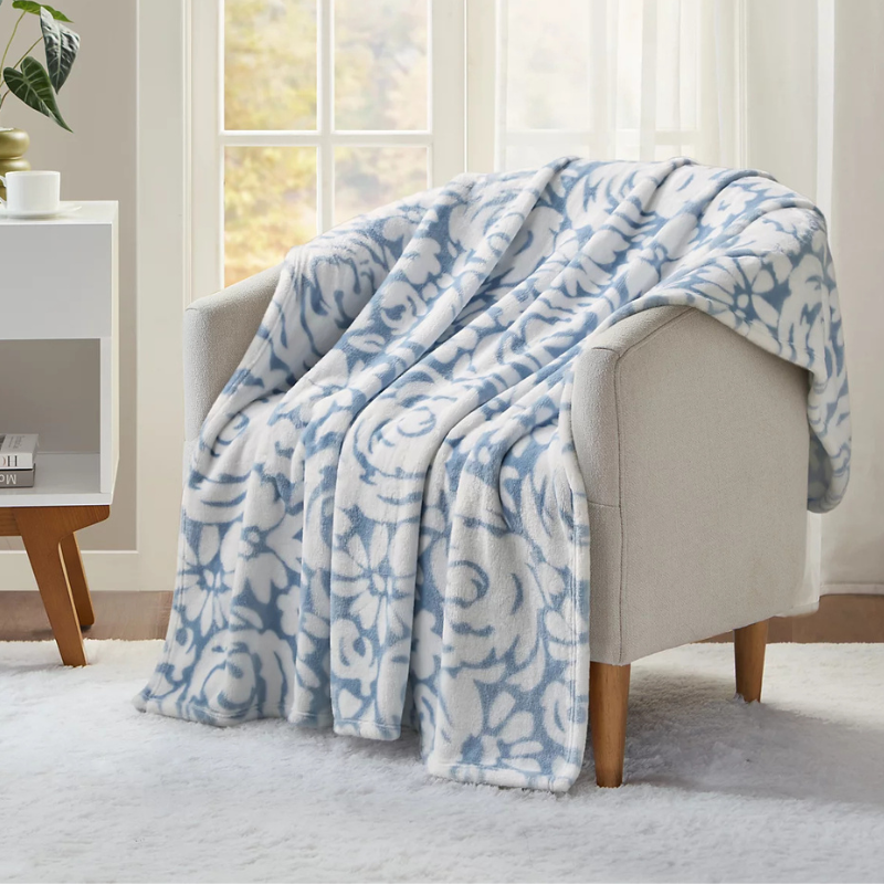 A light gray upholstered chair is draped with a soft, plush blanket featuring a blue and white floral pattern. A small table with a book and potted plant sits nearby, and the setting includes a window and a cozy white rug.