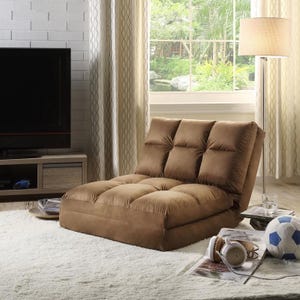 A brown adjustable floor sofa chair with tufted cushions is placed in a cozy living room with a large window, next to a lamp, a side table with books, and items like a soccer ball and headphones on the floor.