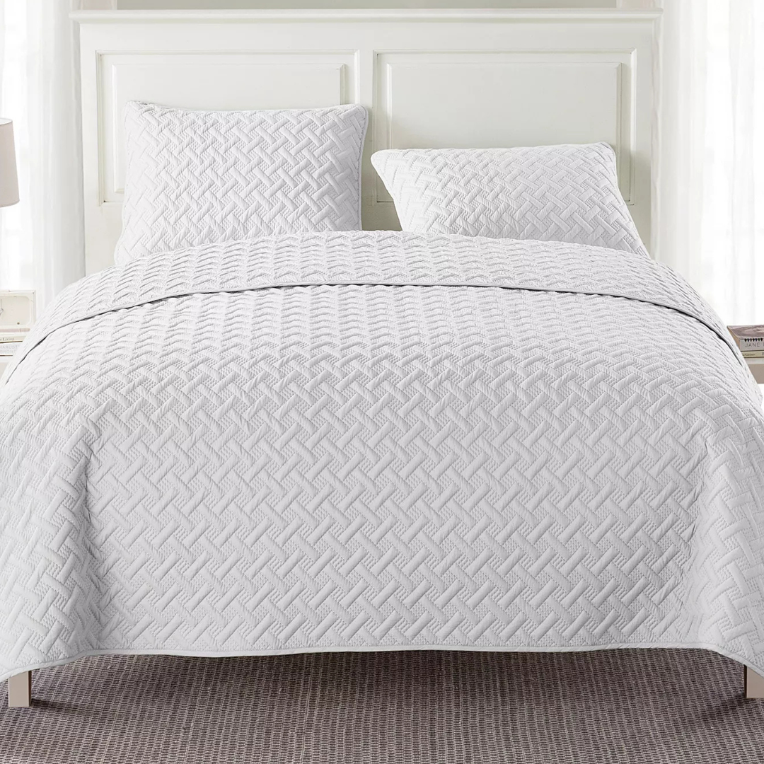 A white quilted bedspread with a textured geometric pattern covers a bed, accompanied by two matching pillows.