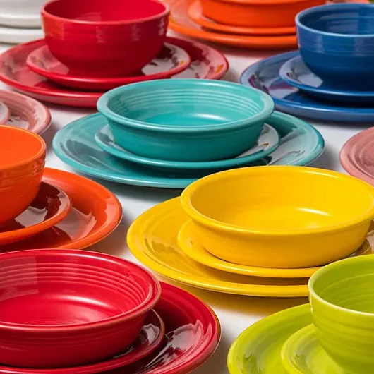 A collection of colorful ceramic dinnerware sets featuring plates and bowls in vibrant red, orange, turquoise, and yellow hues arranged on a white surface.