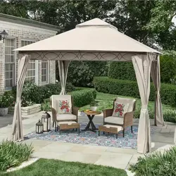 A patio with a beige canopy gazebo features two wicker armchairs with floral cushions, a small round table, and decorative lanterns on a patterned outdoor rug.