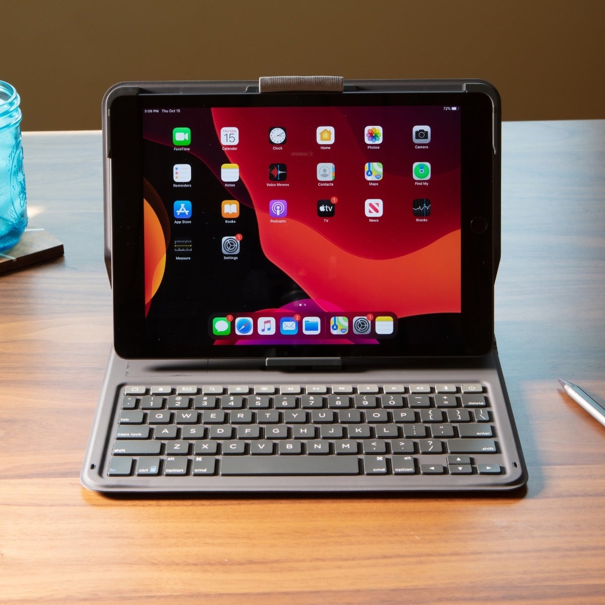 An iPad with a 10.9\“ screen is encased in a Messenger Folio 2, with a black keyboard attached, resting on a wooden surface. The screen displays several app icons, and a pen lies nearby.