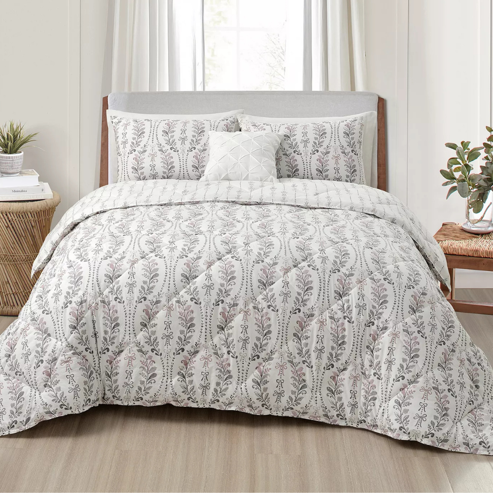 A bed is made up with a comforter and pillow set featuring a floral pattern in soft colors, accompanied by a small decorative pillow. The bedroom includes a wooden nightstand with plants and a stacked book.