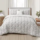 A bed is made up with a comforter and pillow set featuring a floral pattern in soft colors, accompanied by a small decorative pillow. The bedroom includes a wooden nightstand with plants and a stacked book.