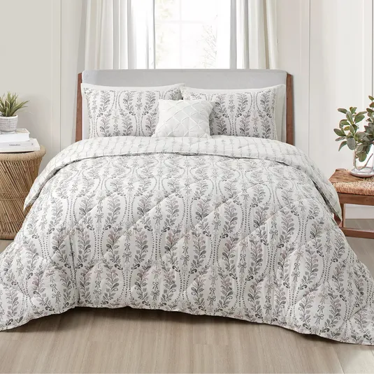 A bed is made up with a comforter and pillow set featuring a floral pattern in soft colors, accompanied by a small decorative pillow. The bedroom includes a wooden nightstand with plants and a stacked book.