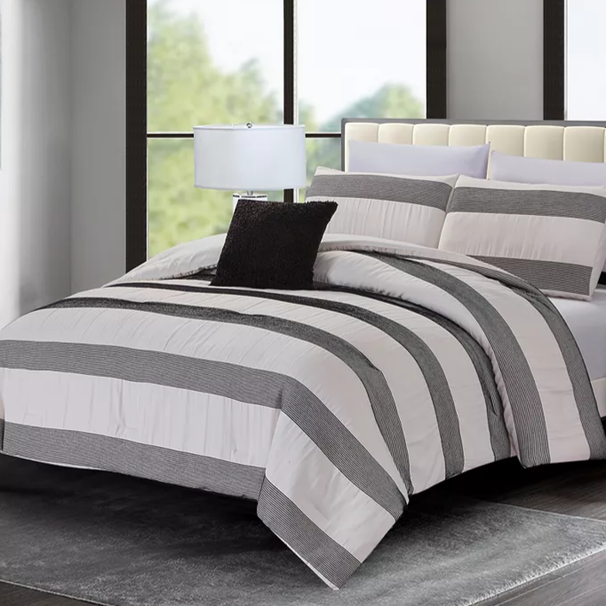 A bedroom features a bed with a striped comforter set in black and white, complemented by matching pillows and a black accent pillow. A table lamp sits beside the bed on a side table, all set against large windows and a plush gray rug.
