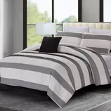 A bedroom features a bed with a striped comforter set in black and white, complemented by matching pillows and a black accent pillow. A table lamp sits beside the bed on a side table, all set against large windows and a plush gray rug.