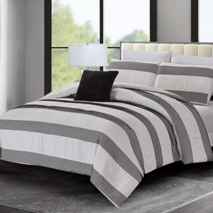 A bedroom features a bed with a striped comforter set in black and white, complemented by matching pillows and a black accent pillow. A table lamp sits beside the bed on a side table, all set against large windows and a plush gray rug.